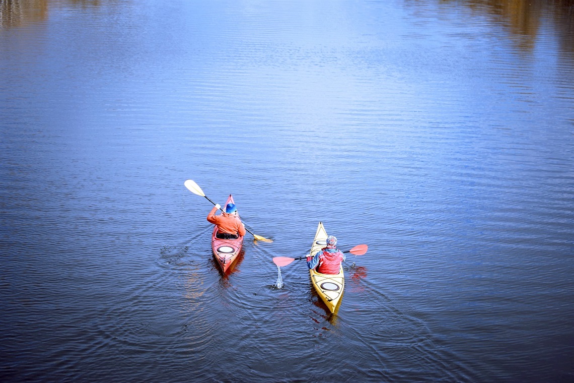 Two kayakers paddle away from the camera in a still body of water.