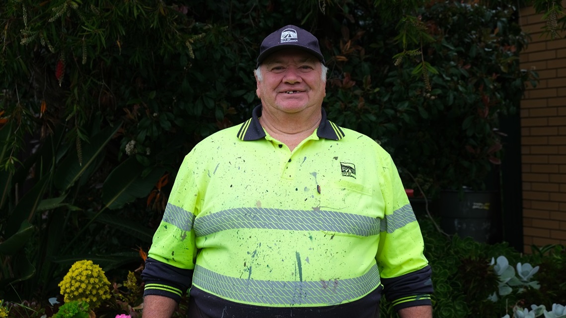 Neville Radford stands in front of some green foliage wearing a high-vis paint-splashed shirt and a cap.