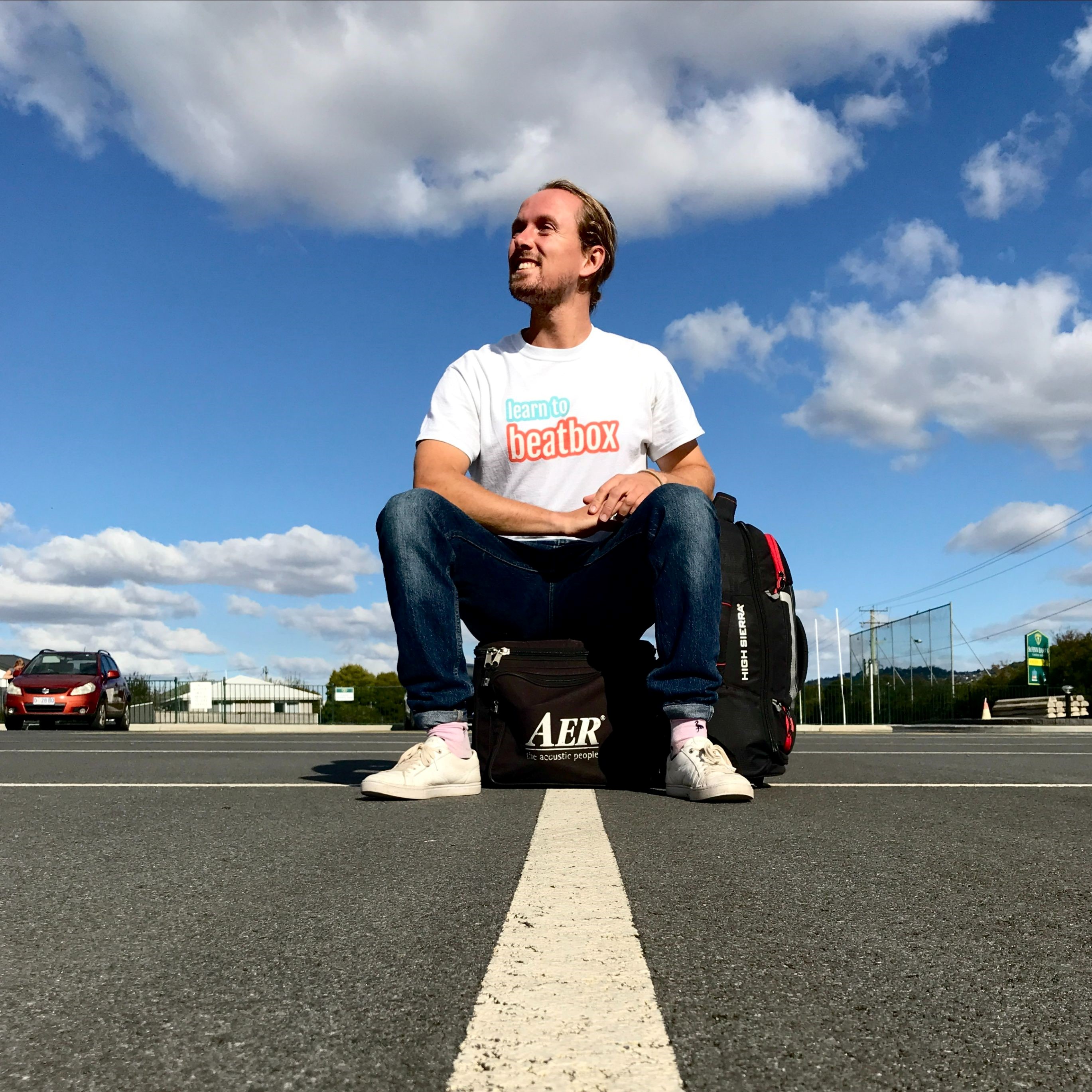 Beatboxing instructor Jobe Burgess sits on a bag smiling in front of a blue sky.