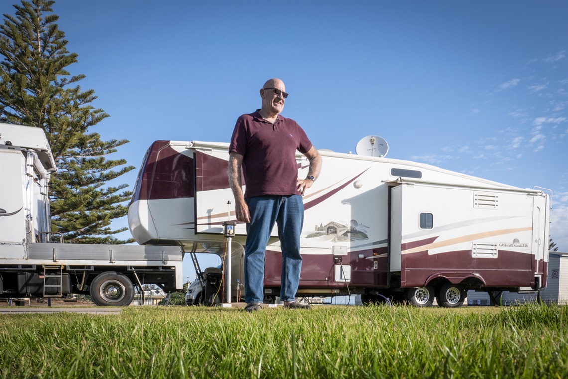 A bald man in blue jeans and a maroon shirt stands on grass in front of his maroon-and-white caravan on a sunny blue-sky day.