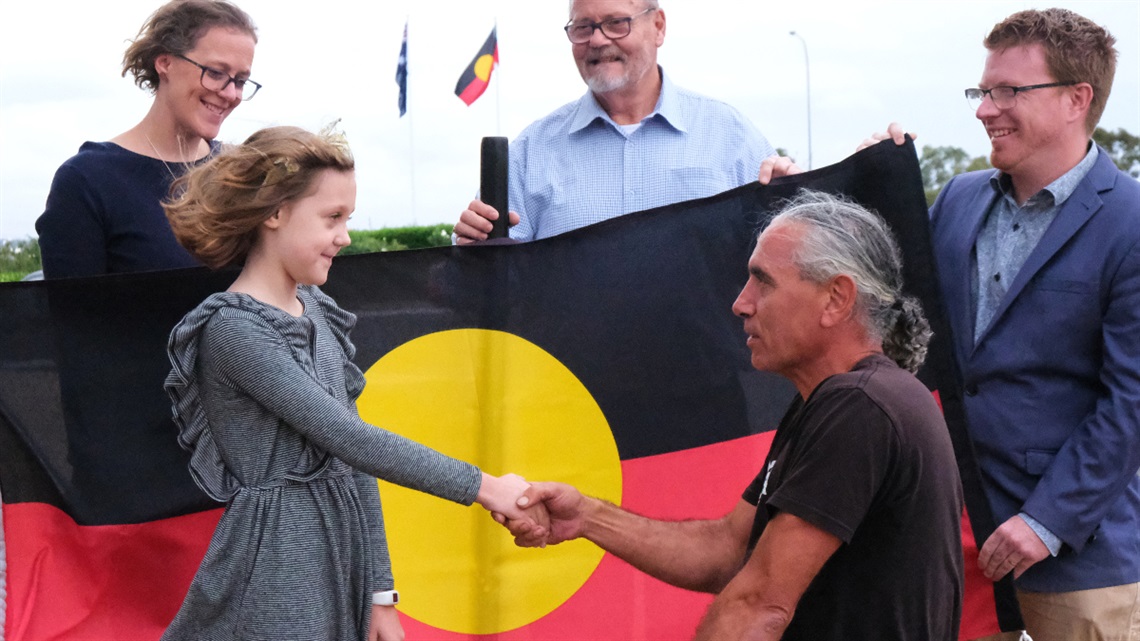 Eight-year-old Aberfoyle Park student Summer shakes hands with Traditional Owner Karl Telfer in front of an Aboriginal flag.