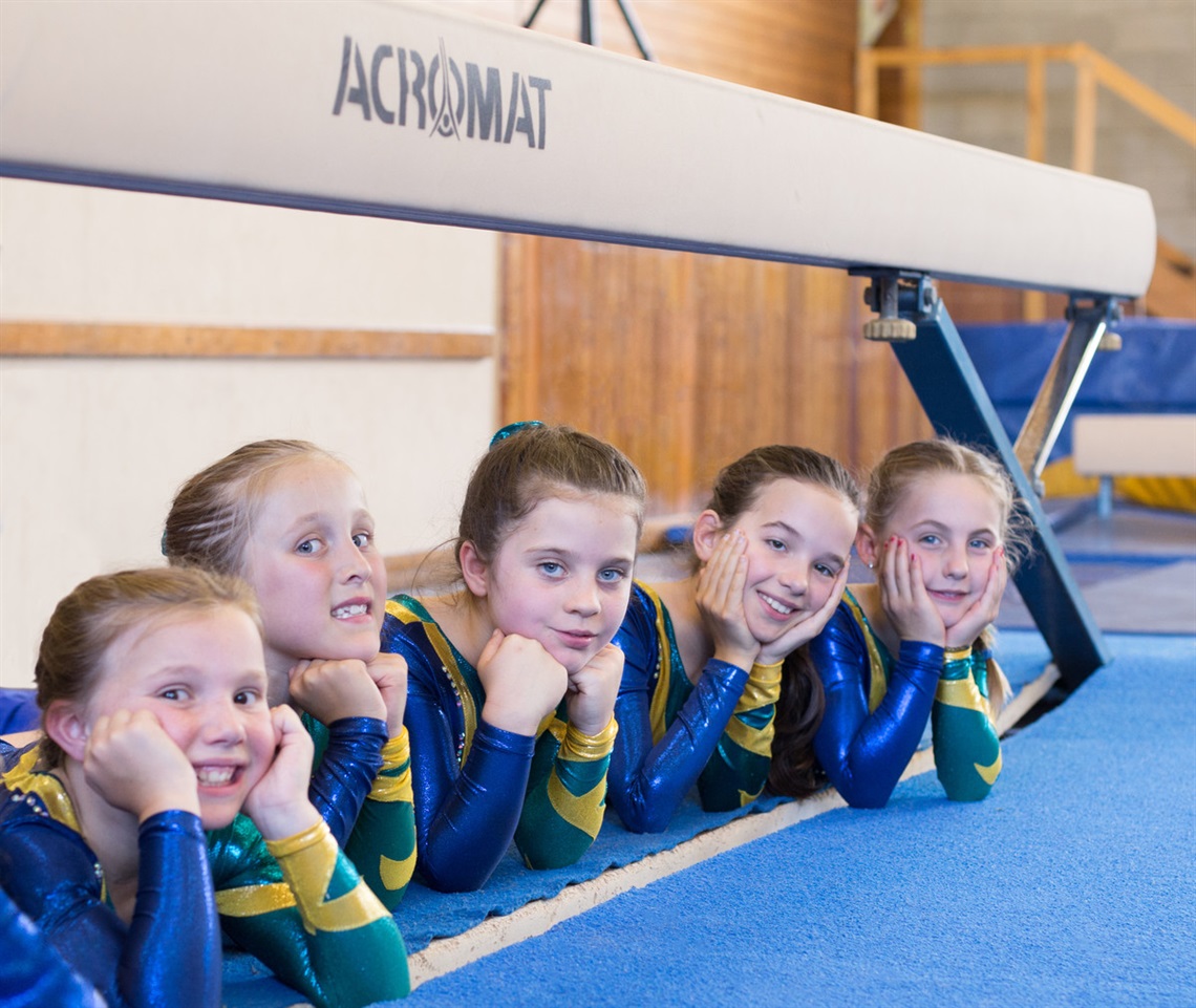 A row of young female gymnasts lying on the floor alongside gym equipment smile for the camera.