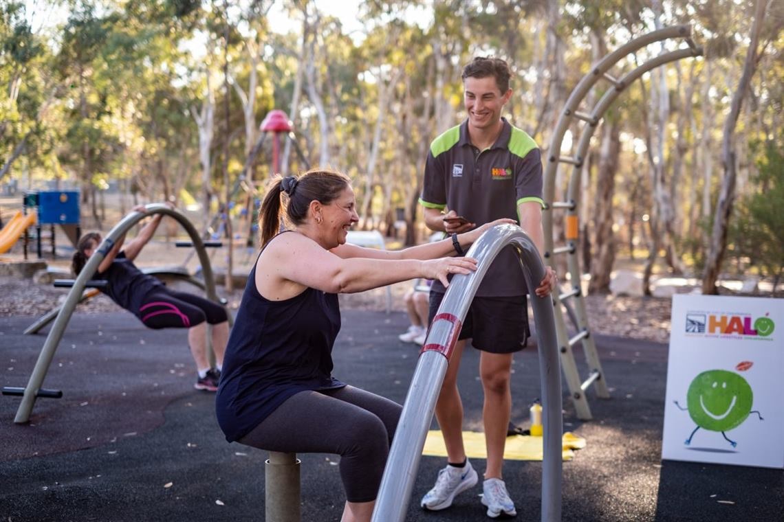 A smiling woman in black workout gear uses outdoor fitness equipment while a polo-shirted personal trainer looks on.