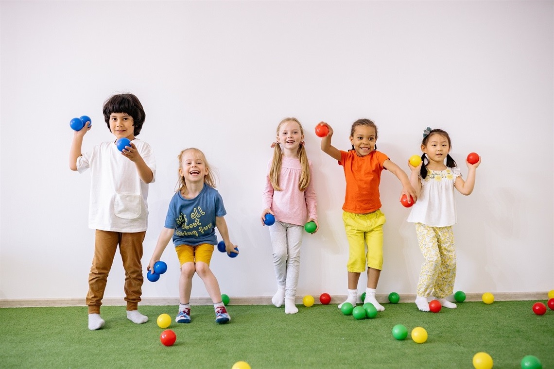 A line of smiling young children post with colourful balls atop artificial turf.