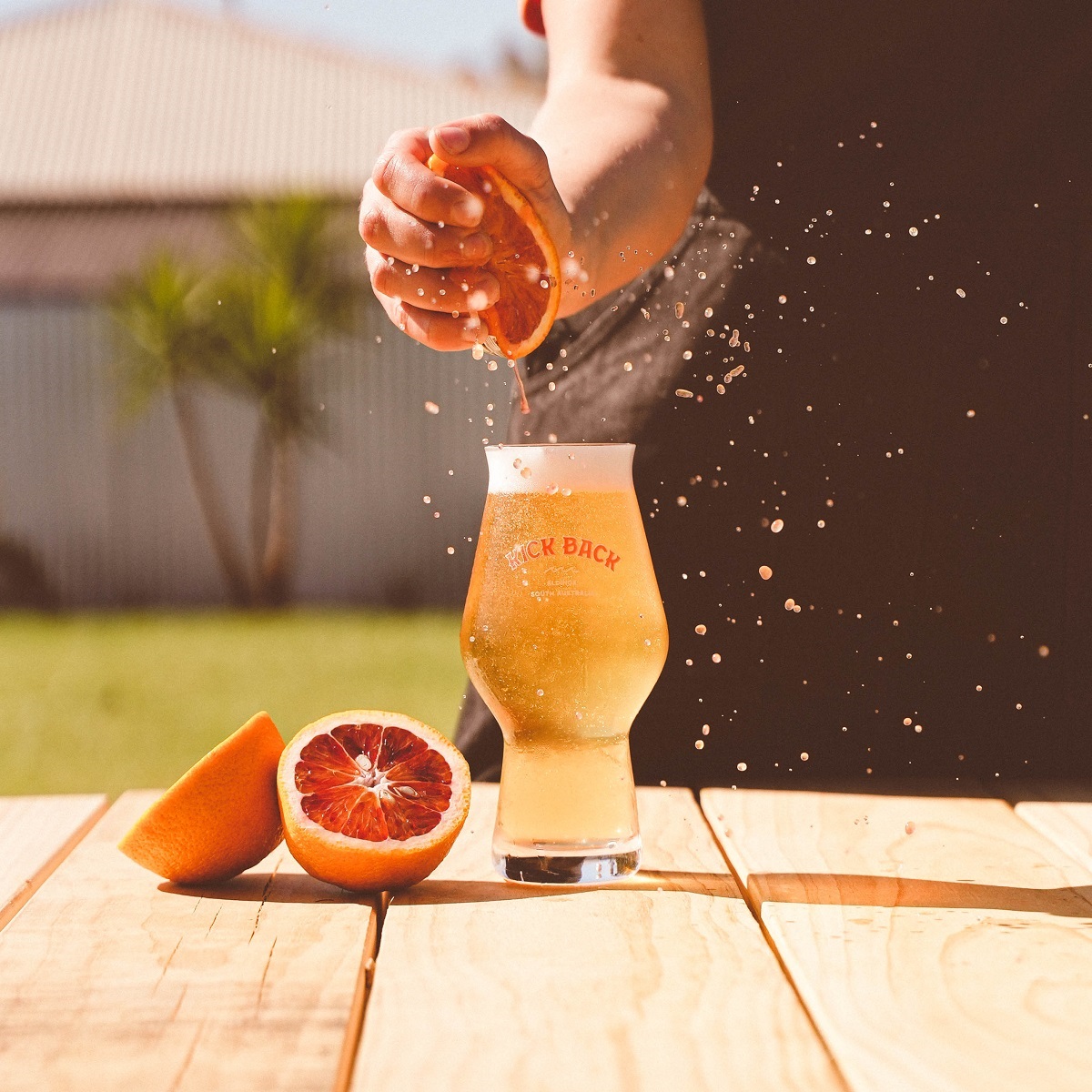 A hand squeezes a blood orange into a cold beer glass atop a timber table.