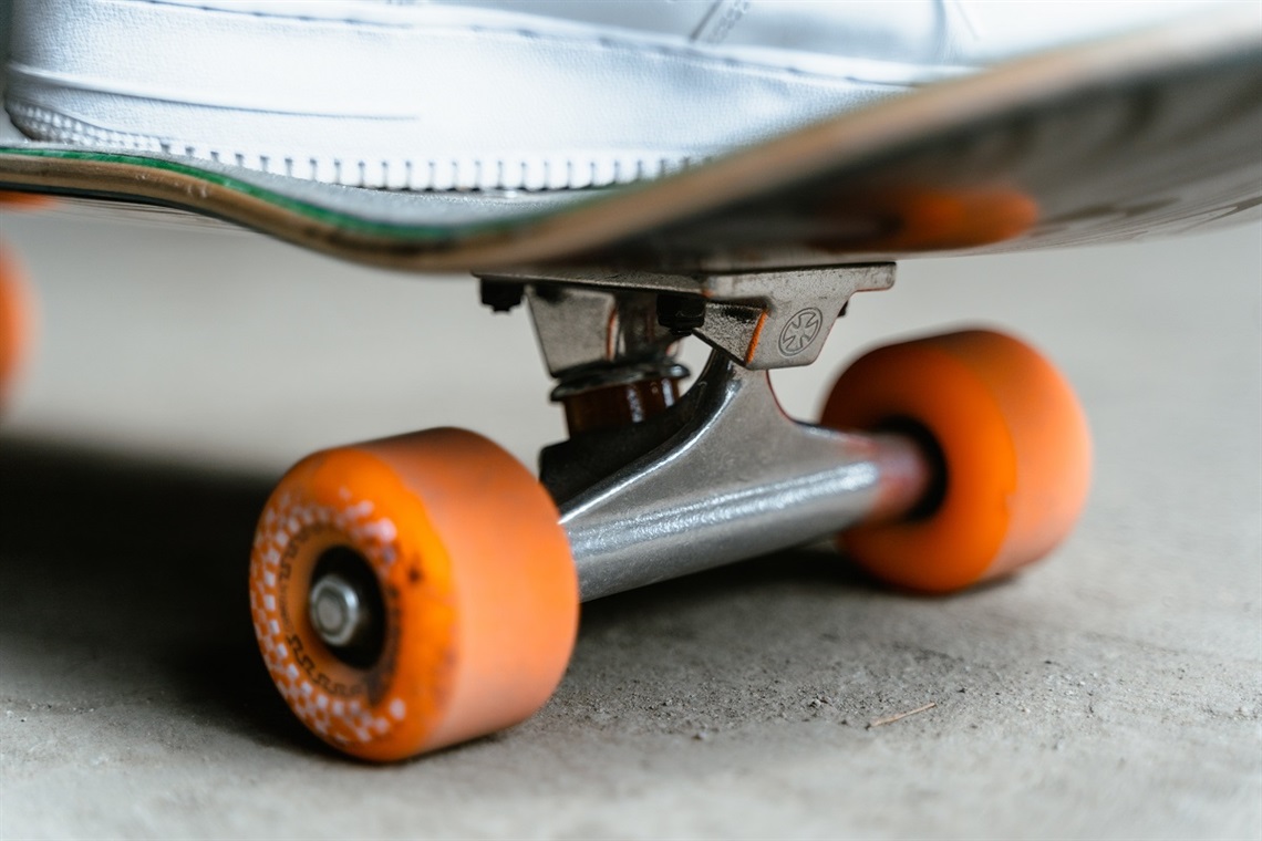 A close-up shot of the orange back wheels and trucks of a skateboard with the sole of a white shoe on top.