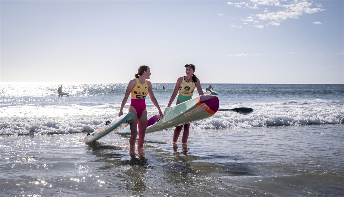 Two young surf life savers smile while carrying their boards from the water.