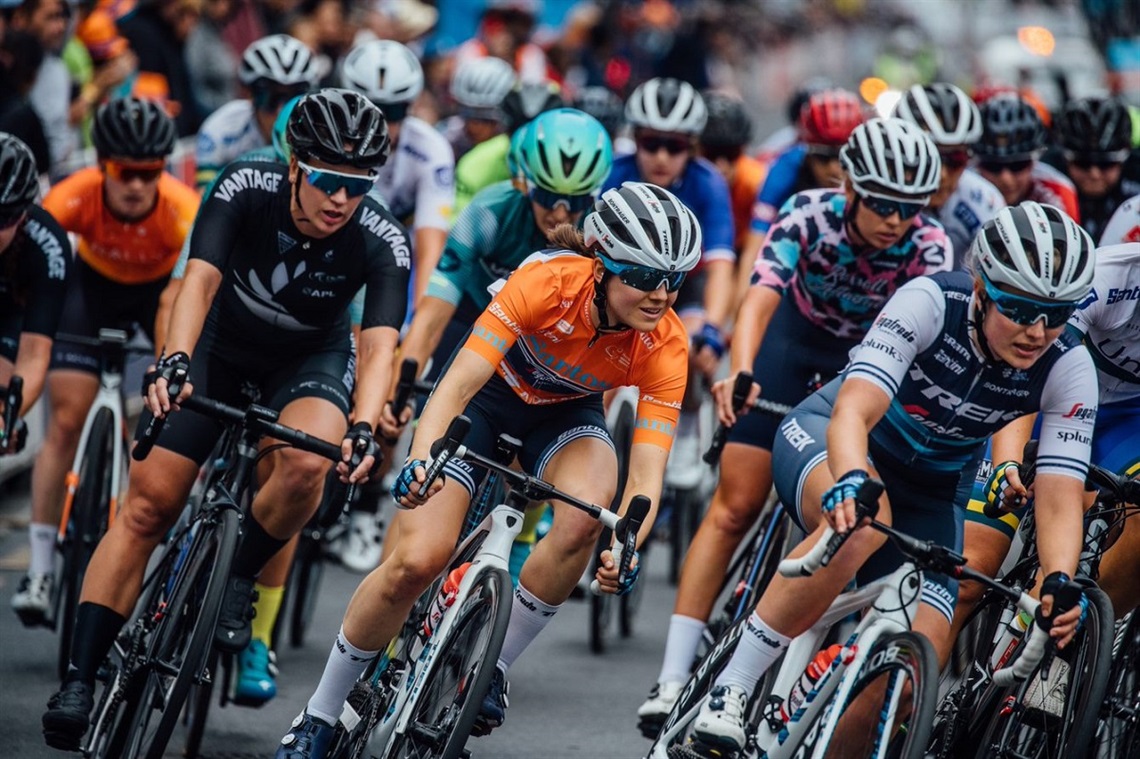 A close-up shot of a pack of women cyclists wearing colourful race attire and sunglasses turning a corner on a grey road.