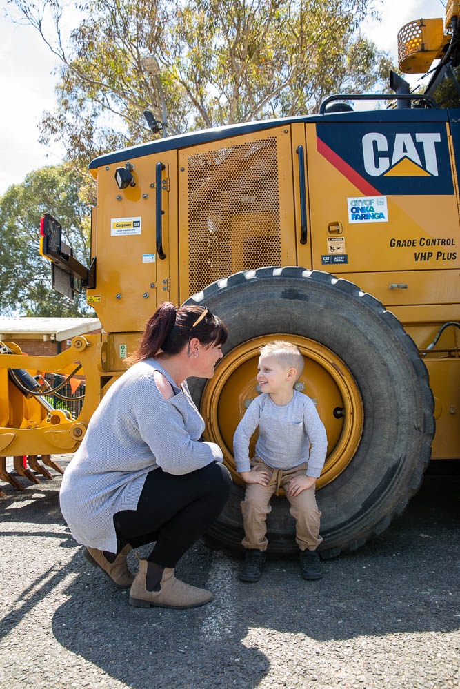 A smiling boy sits inside the wheel of a yellow council construction vehicle alongside his mum.
