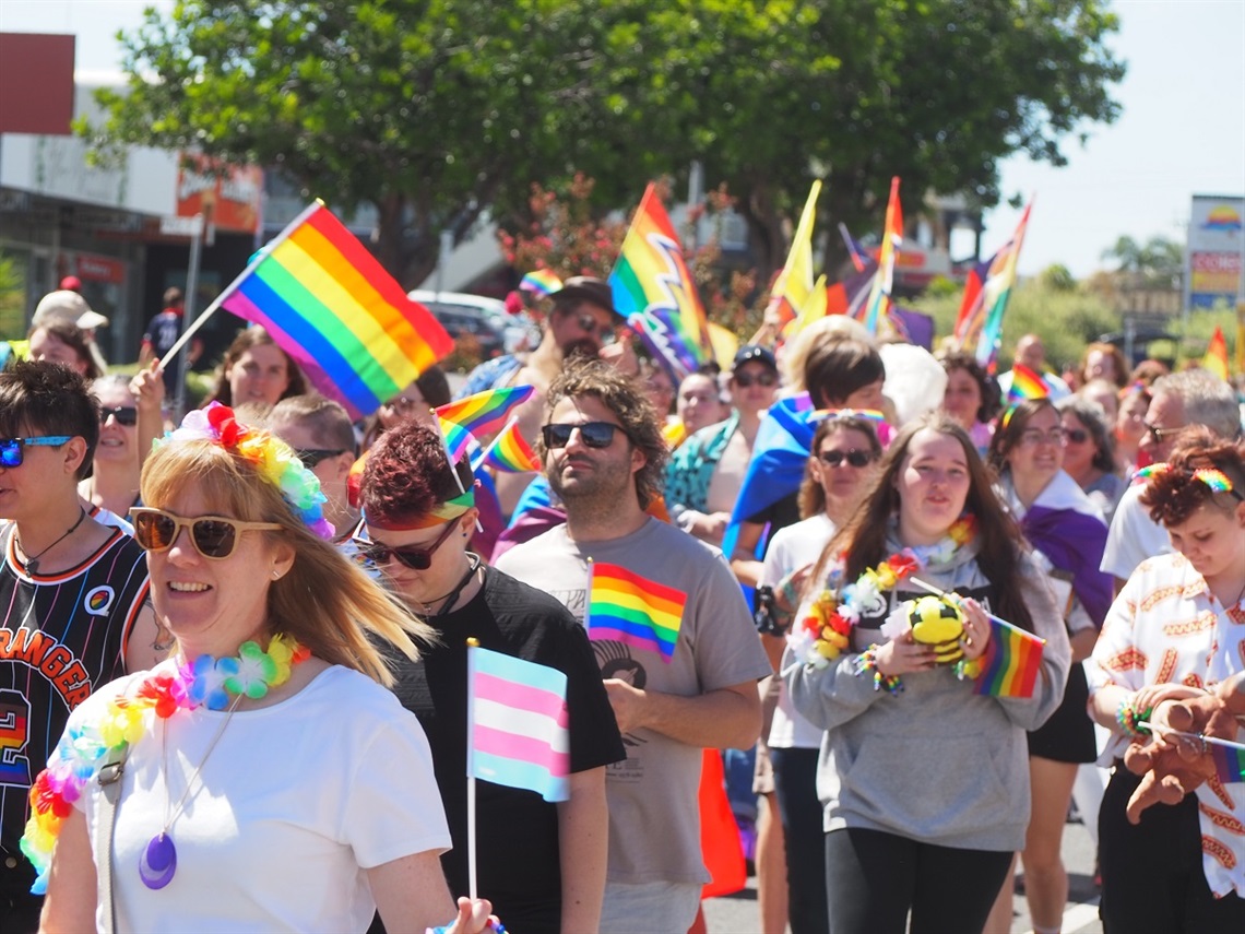 Crowds of people waving rainbow flags walk towards the camera along Beach Road, Christies Beach.