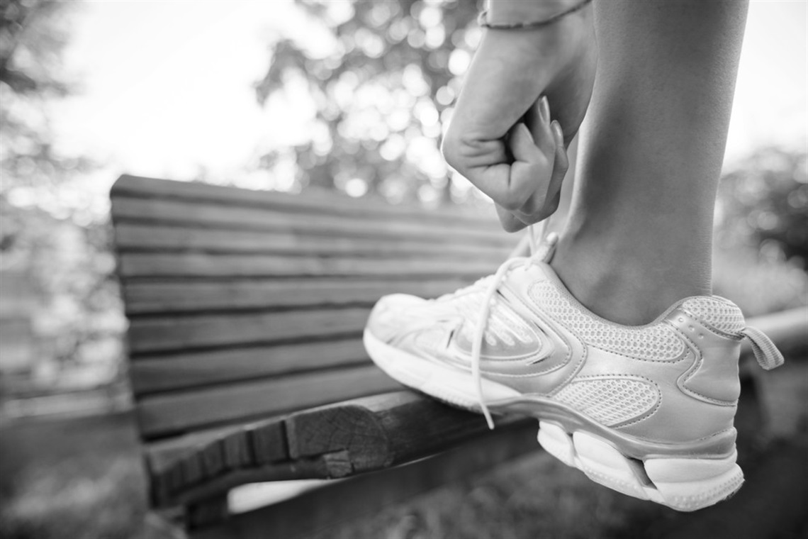 A close up black-and-white photograph of someone lacing up their white walking or running shoe, which is resting on a wooden park bench, with trees and sunlight in the background.