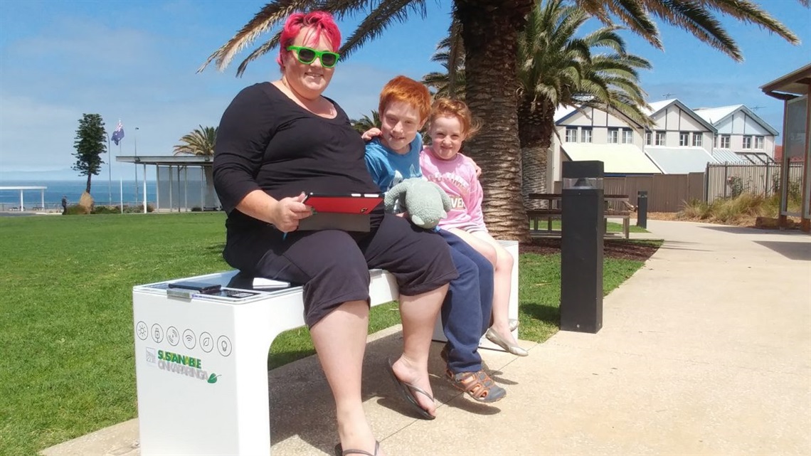 RESIDENT AMY HERMANN, PICTURED WITH HER CHILDREN, USING THE NEW SMART BENCH TO CHARGE HER MOBILE PHONE.
