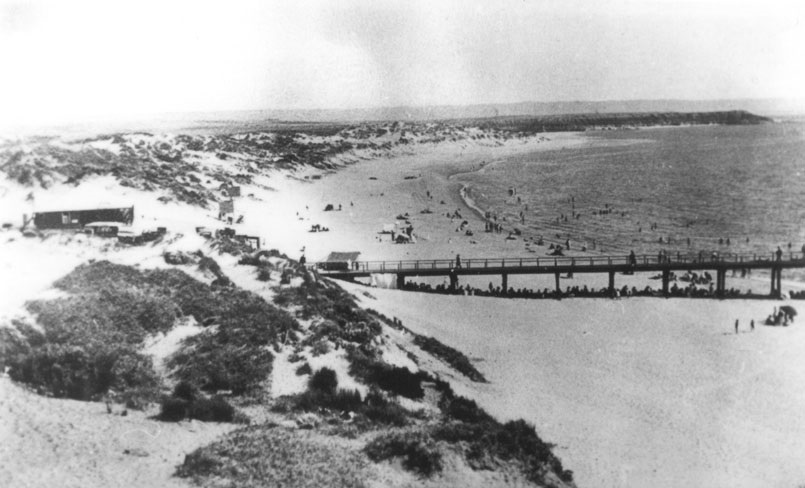 A black and white view from Witton Bluff, looking down towards the Port Noarlunga jetty on a busy beach day fringed by sand dunes.