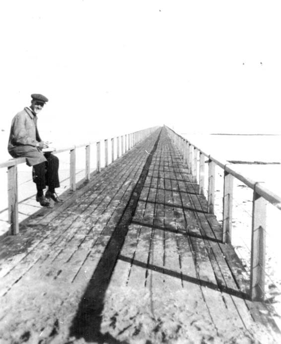 A black and white photo of a smiling man sitting on the railing of the Port Noarlunga jetty in the 1920s.