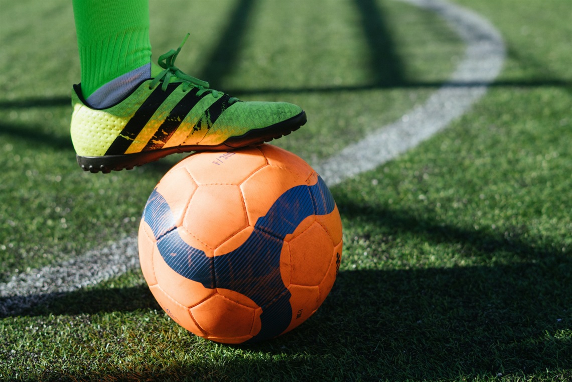 A soccer player's green and black boot sits atop an orange and blue ball on a grass lined pitch.