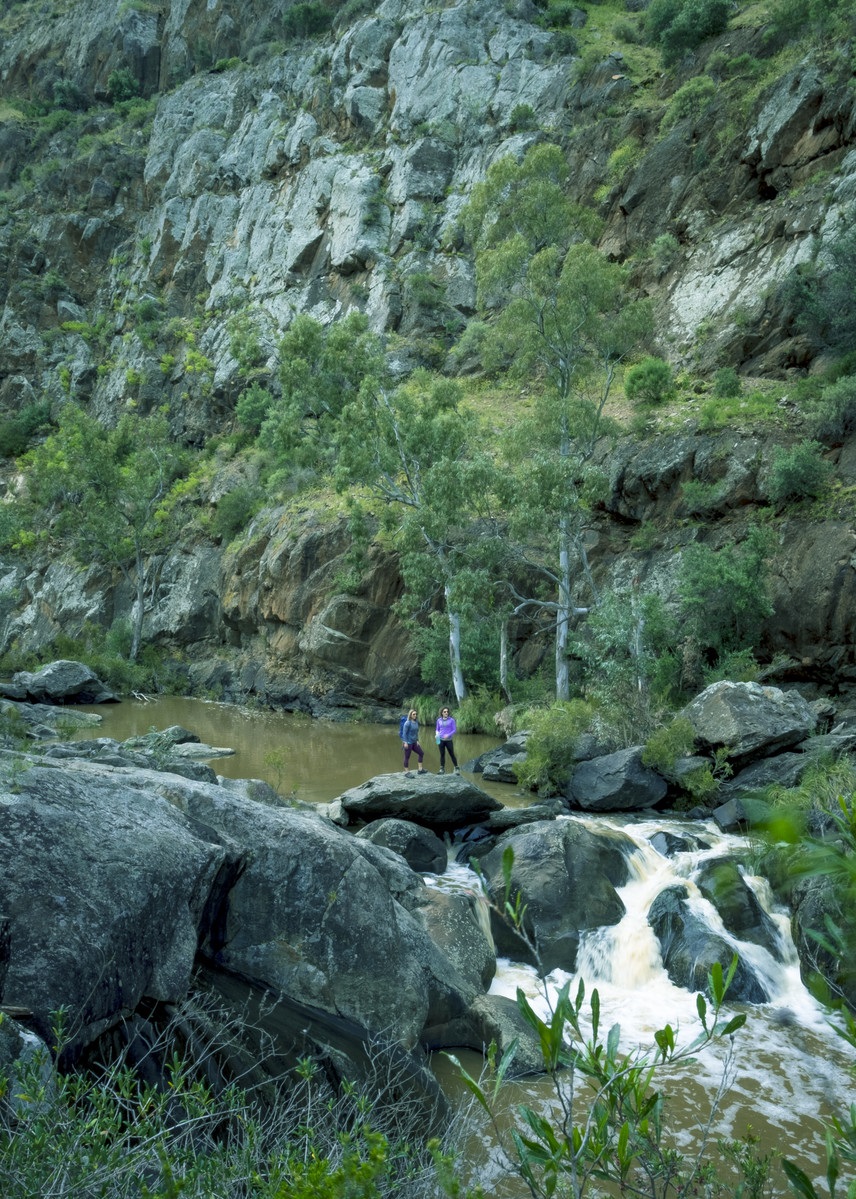 A pulled-back shot of two walkers standing on some rocks alongside some tall cliffs and a waterfall.