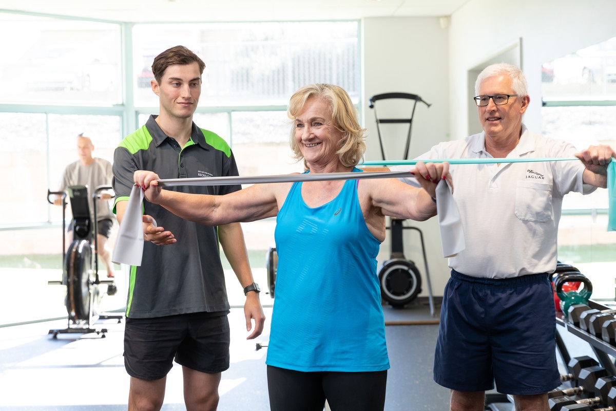 Two smiling gym goers use elastic bands to work out alongside the watchful eye of a personal trainer.