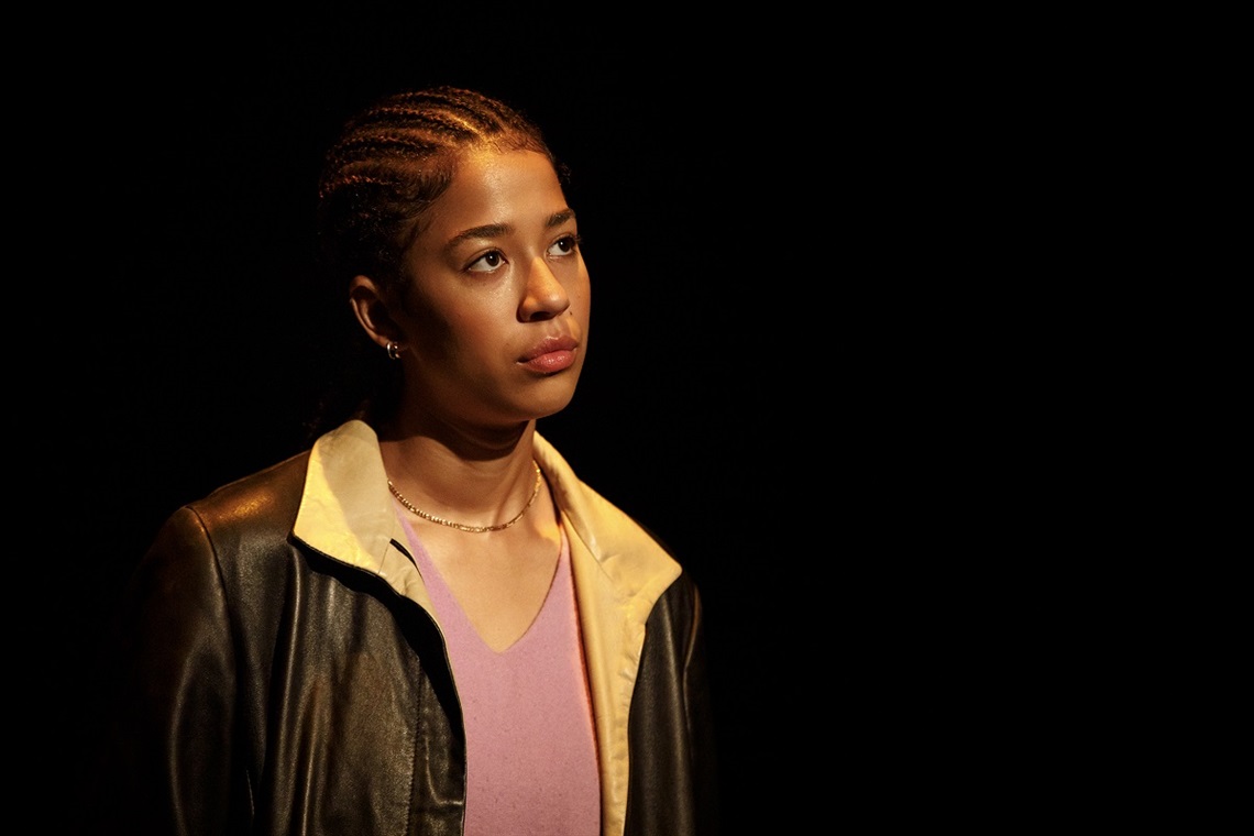 A young woman stares away from the camera on a spotlighted stage surrounded by darkness.