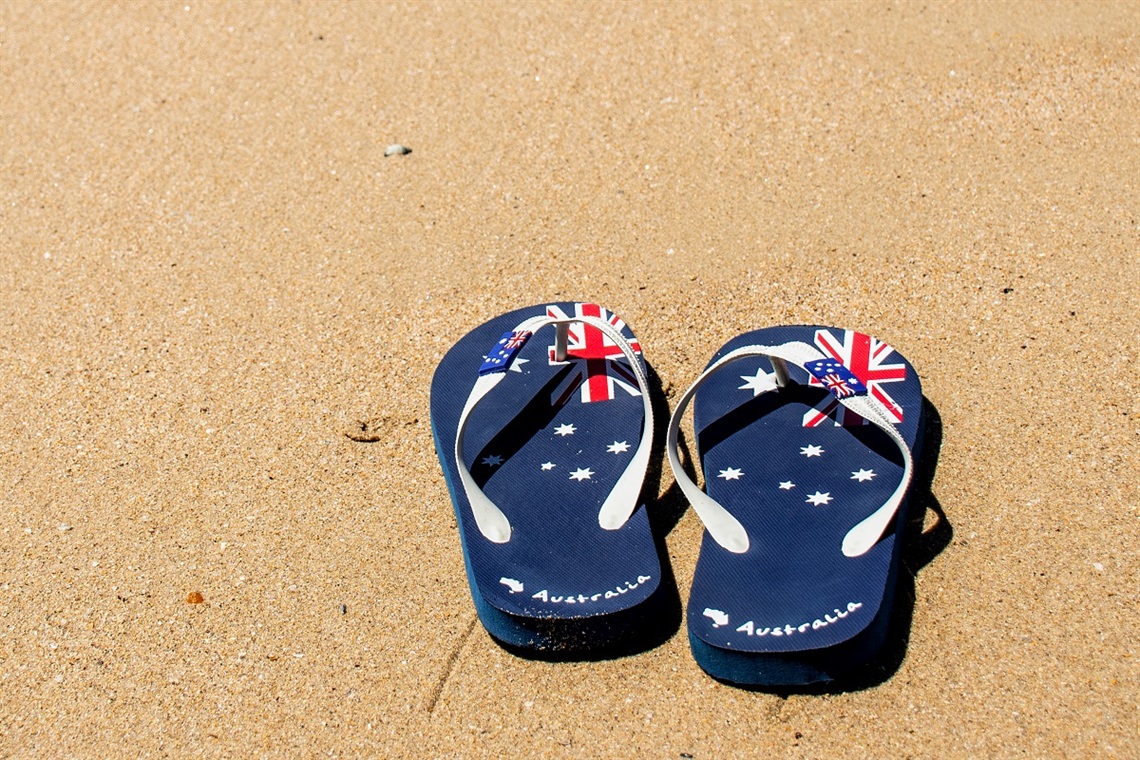 A pair of blue and white thongs featuring the Australian flag design sit on the sand.