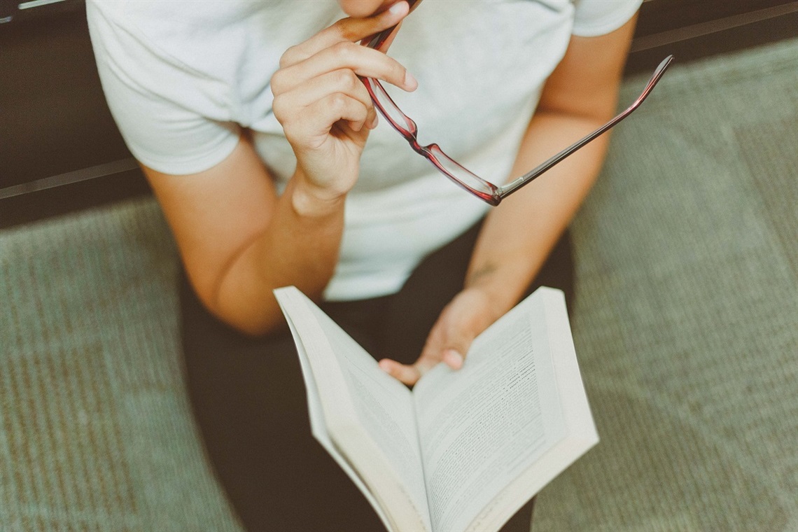 A person, their head out of frame, holds a pair of glasses and a blurred, open book.