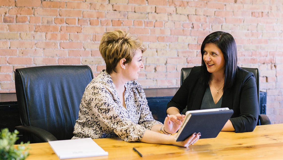 Two women in a corporate setting have a discussion at an office table.