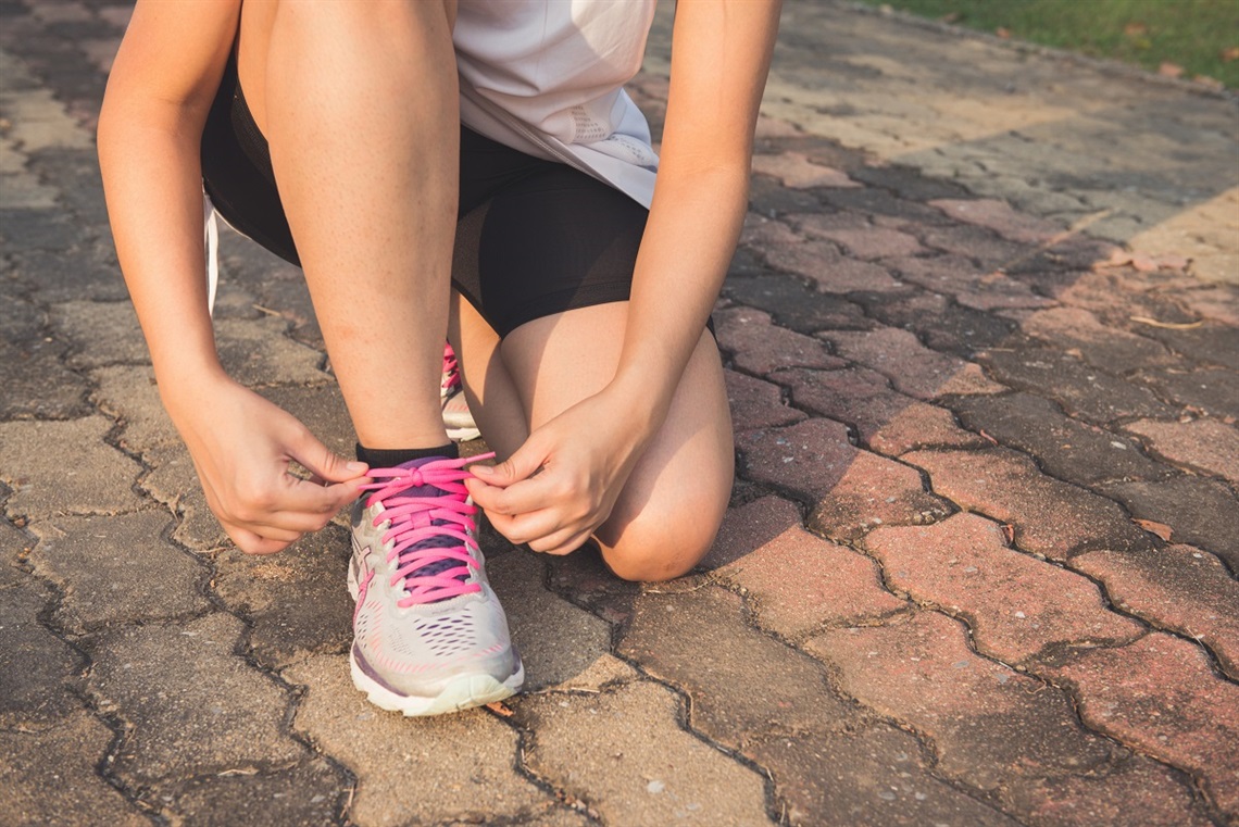 A person on one knee ties the pink laces of their grey running shoe on a brick path.