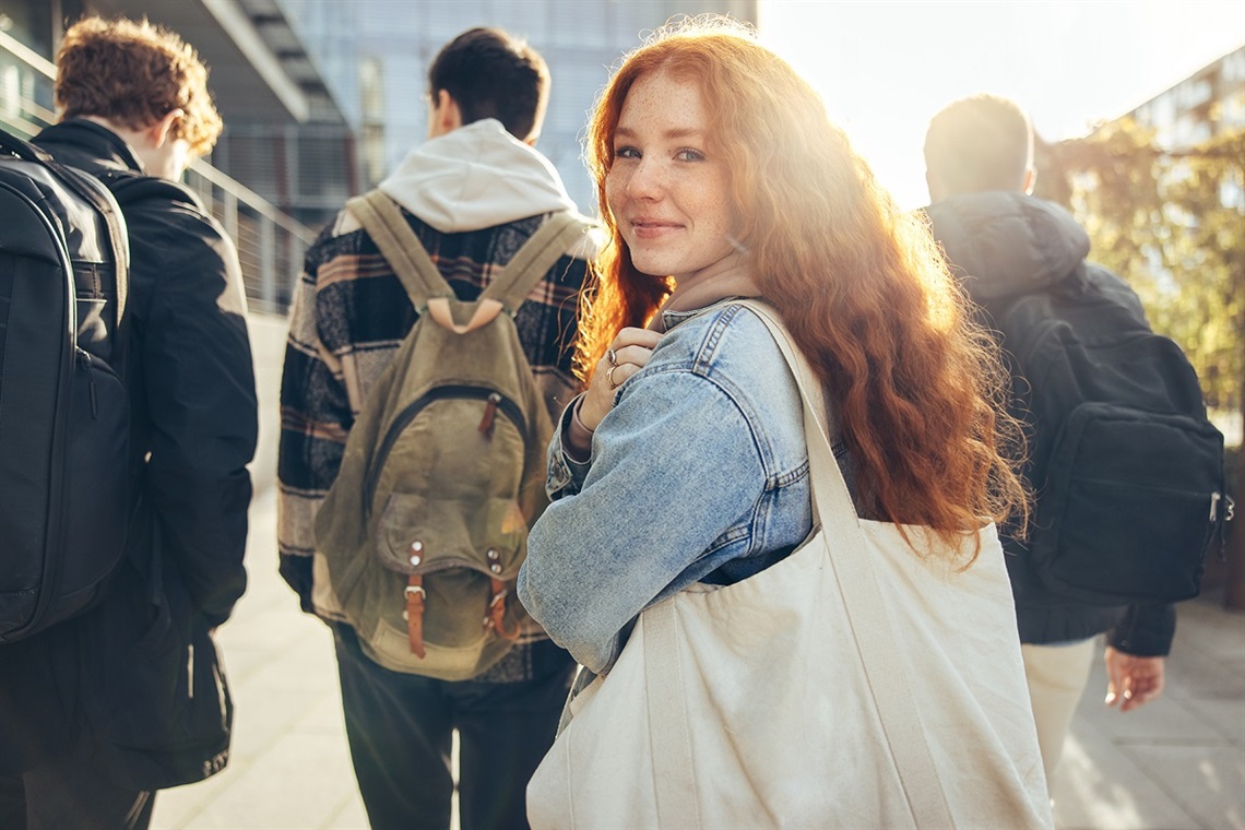 A young fair-skinned red-haired woman wearing a denim jacket turns and smiles at the camera.