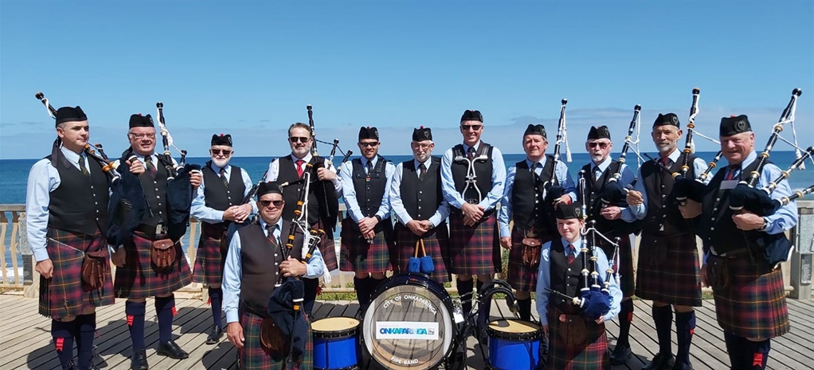 13 pipe band members in kilts pose with their bagpipes and drums.