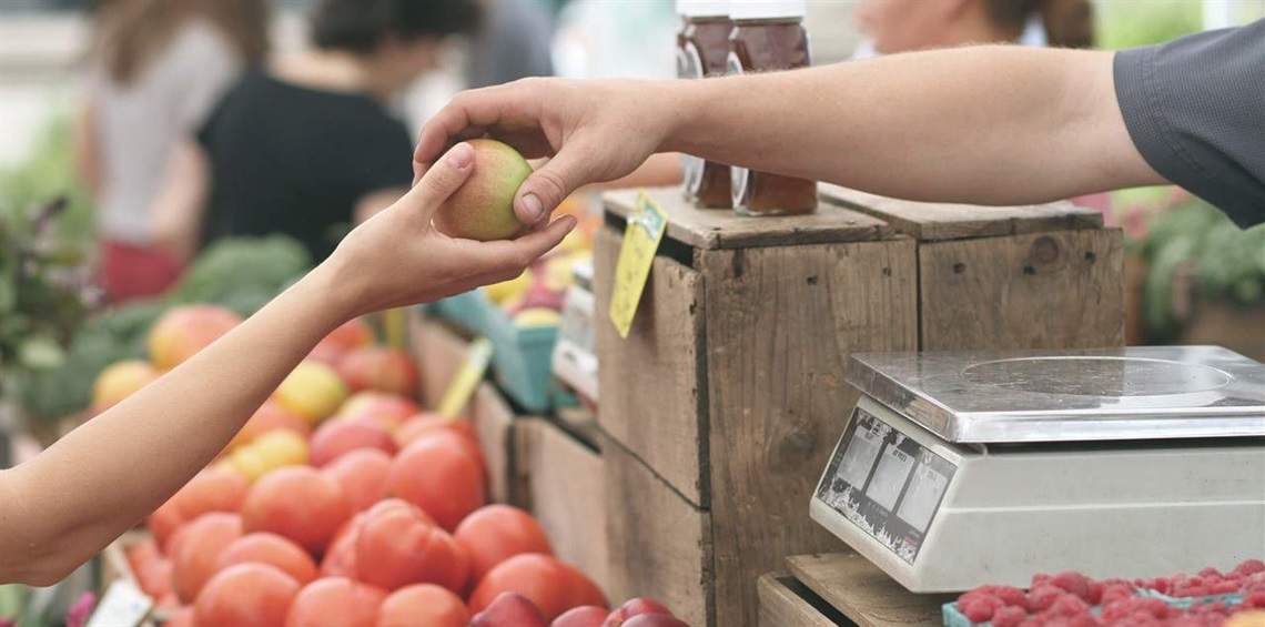 A hand passes an apple to a shopkeeper at a fruit-filled grocery stand.