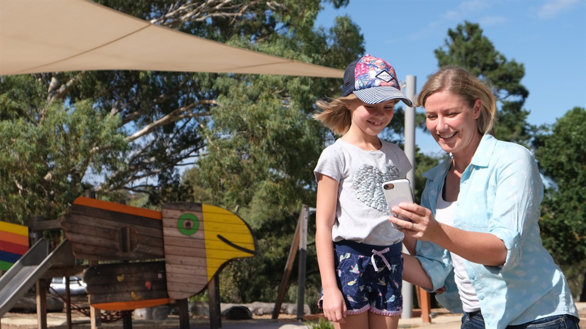 A mother and daughter smile while looking at a phone at Thalassa Park's playground.