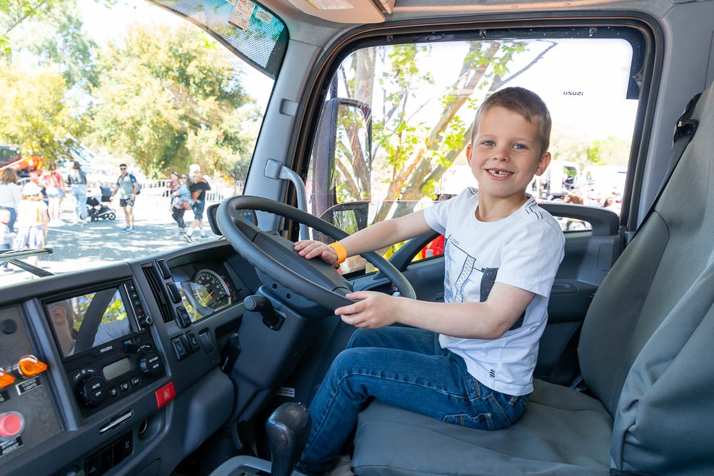 A smiling boys sits inside a council truck, hands on the steering wheel.