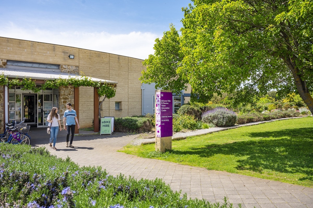 A man and woman walk from the McLaren Vale visitor information centre on a clear and sunny day alongside lush green grass, a large tree and purple flowers.
