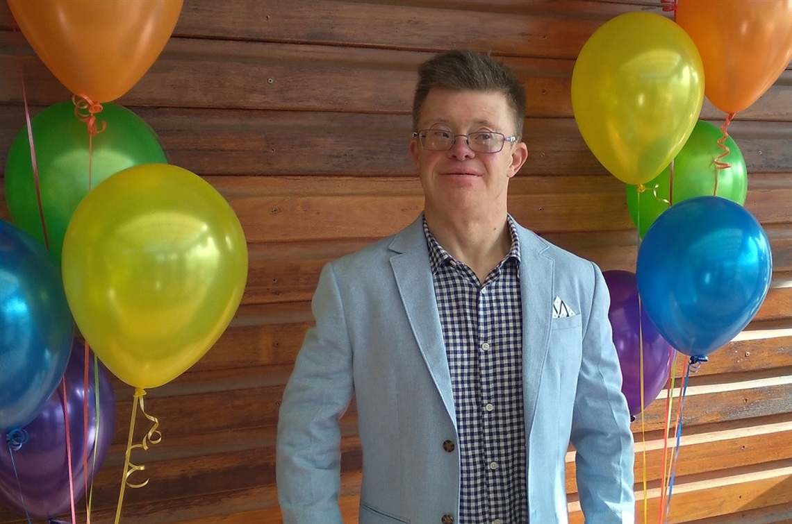 Ben Wishart in a dapper blue suit stands alongside two sets of colouful balloons.