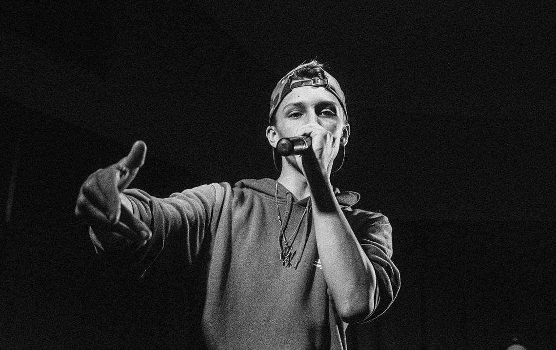 A black-and-white photograph of a young man with his cap backwards and wearing a hoodie, rapping into a microphone with his hand outstretched towards the camera.