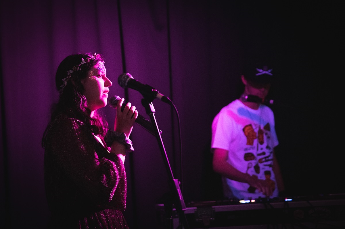 A young woman bathed in purple light on stage sings into a microphone with a keyboardist in the background.
