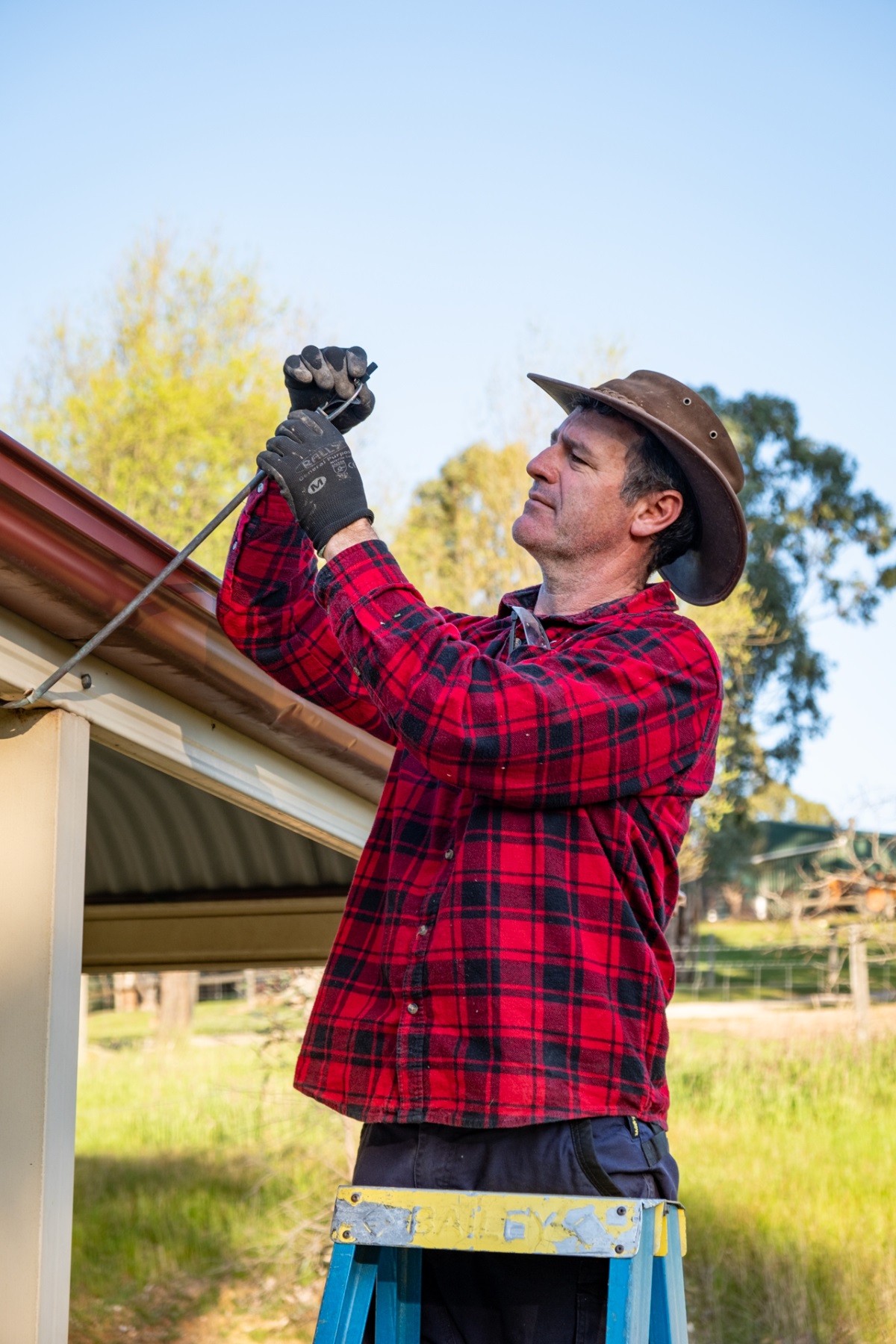 Andrew Ayles, wearing a red and black flannelette shirt, a brown hat and gloves, stands atop a ladder and checks bushfire sprinklers near his gutters.