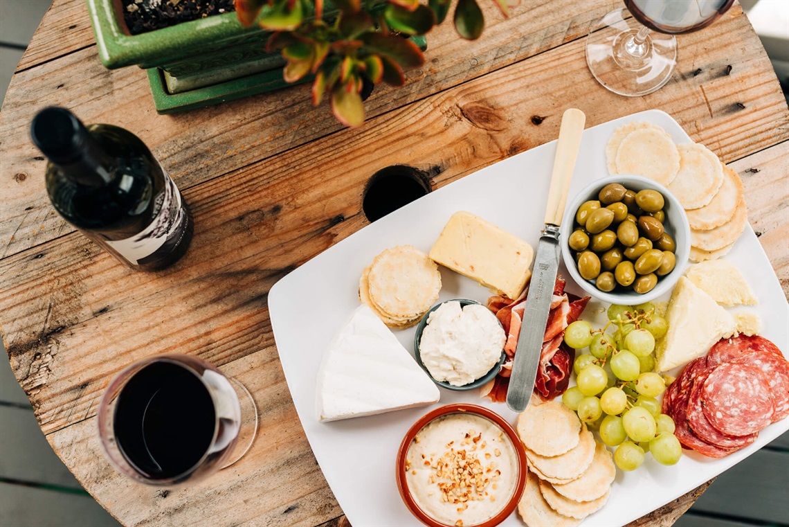 A wooden tabletop with a cheese platter, a bottle and a glass of wine on top.