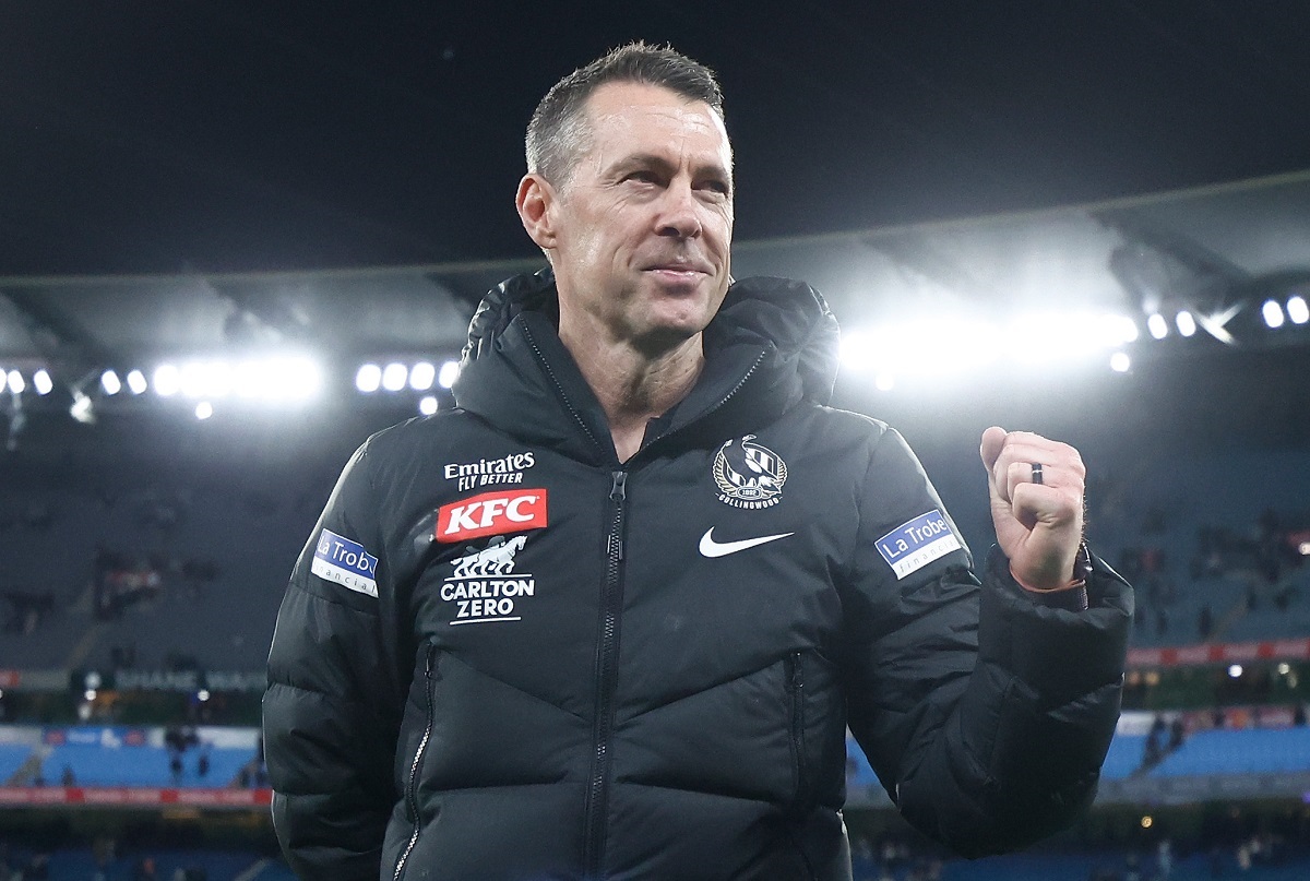 Collingwood Football Club coach Craig McRae smiles and clenches his fist while standing on an oval at night.