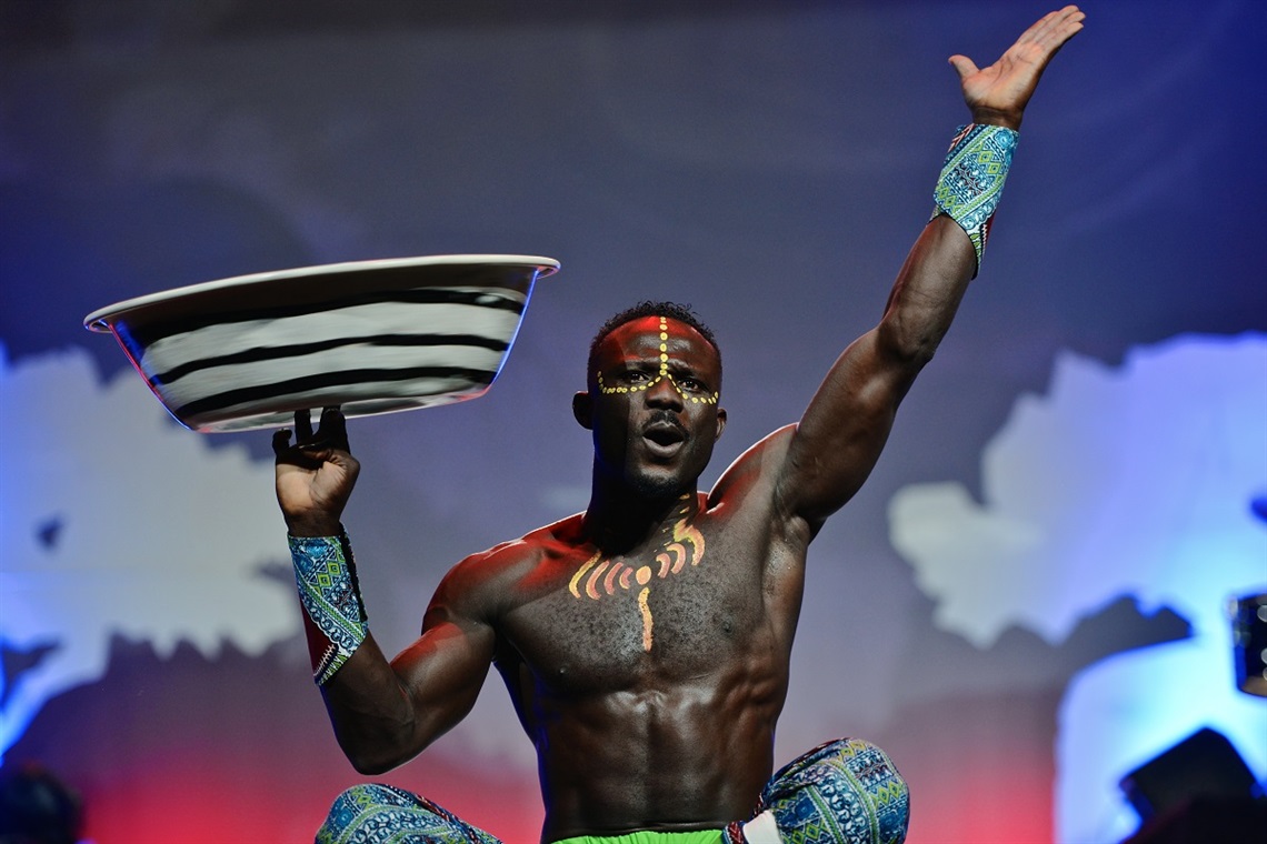 A shirtless muscular performer with a painted face and chest balances a large bowl on his fingers.