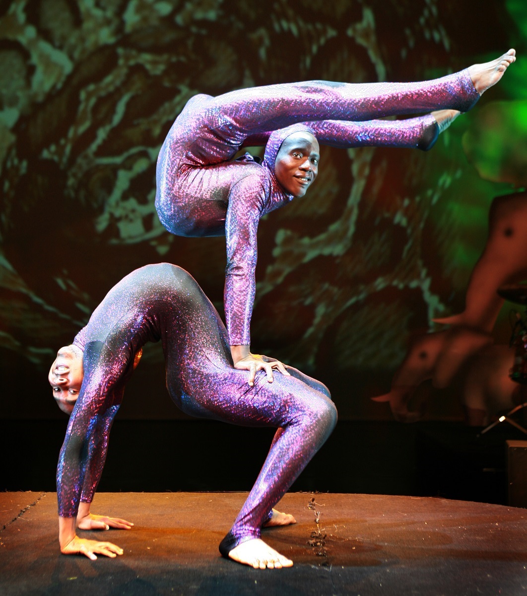 Two performers wearing sparkly purple jumpsuits balance off one another in a gymnastic pose.