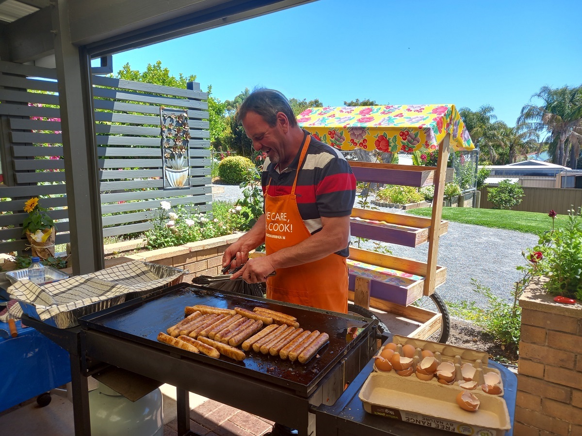 A smiling man wearing an orange apron cooks sausages on a barbecue.