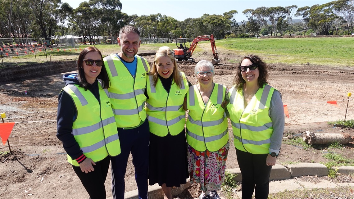 Representatives involved with the Knox Park multi-use athletics facility smile for the camera in high-vis vests as work takes place with a digger behind them.