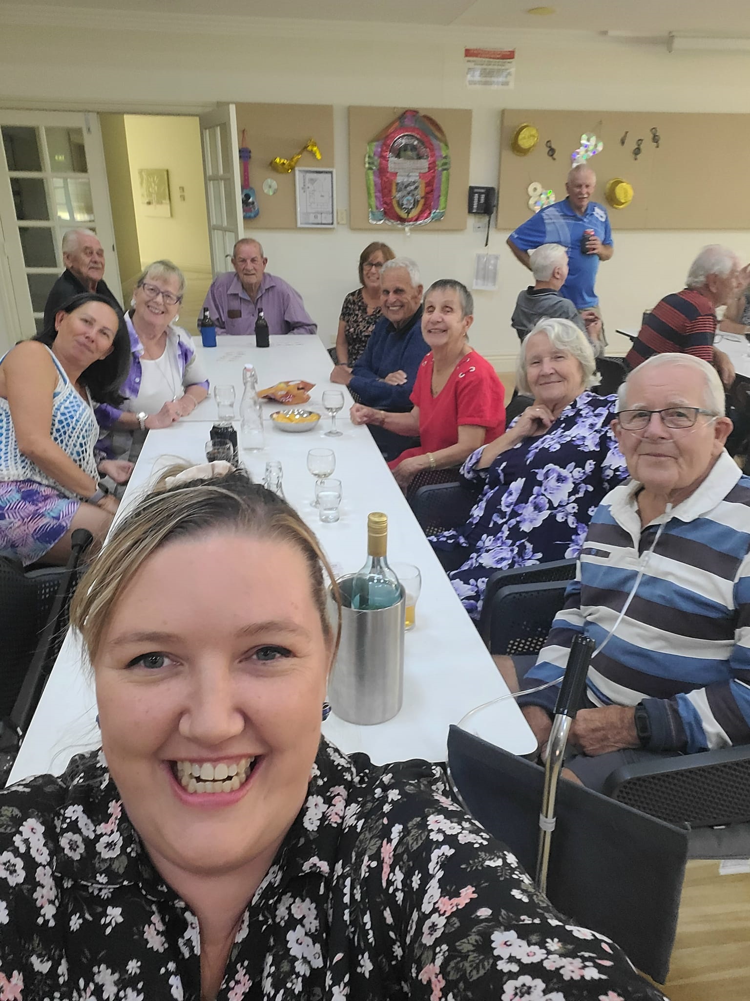 Lauren Jew smiles in a selfie with a community group seated at a table behind her.