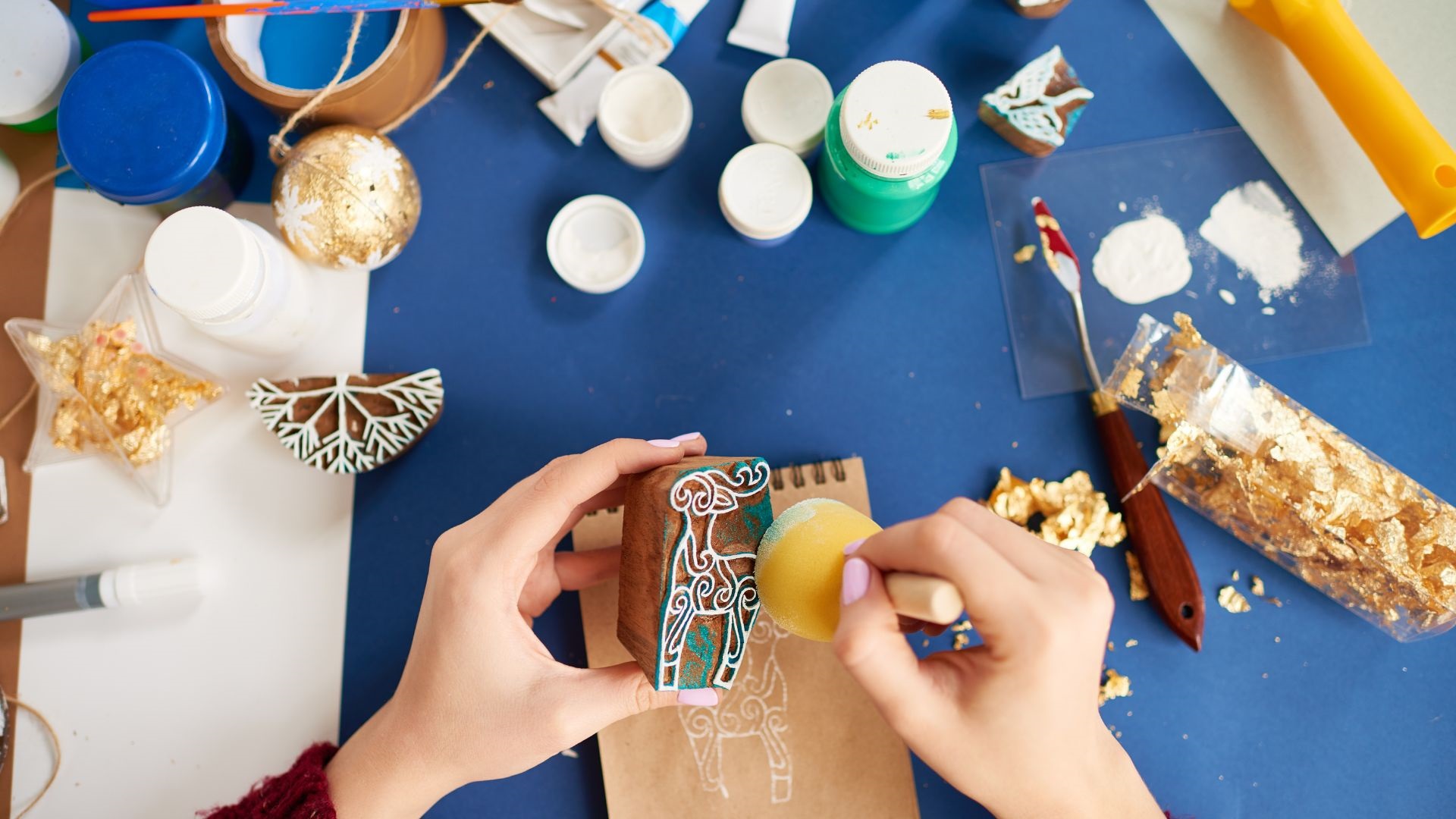 A close-up photo of an artist's hands applying paint to a carving block with paints, glues and other crafty objects on a blue tabletop in the background.