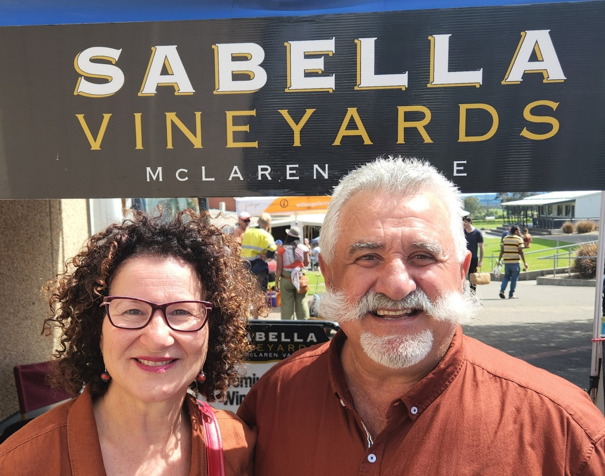 Councillor Marisa Bell smiles alongside a moustachioed business owner and a sign reading 'Sabella Vineyards, McLaren Vale'.