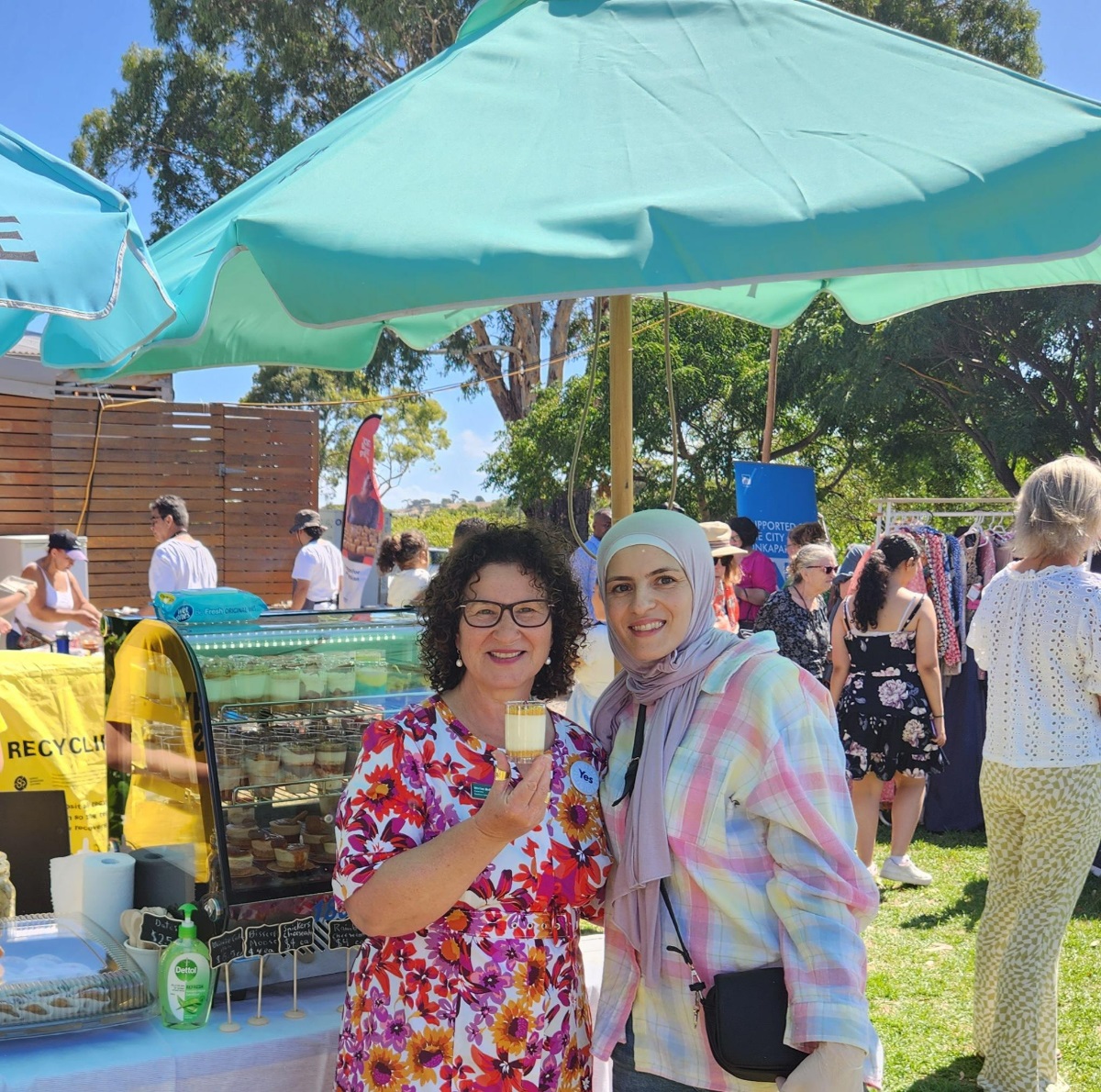 Councillor Marisa Bell smiles and holds a dessert alongside a resident wearing a headscarf under an umbrella.