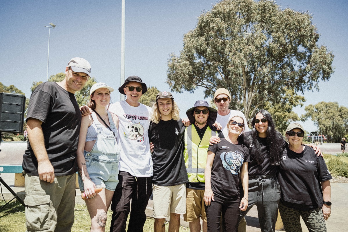 A group of people at a skateboarding event smile at the camera with their arms around one another.