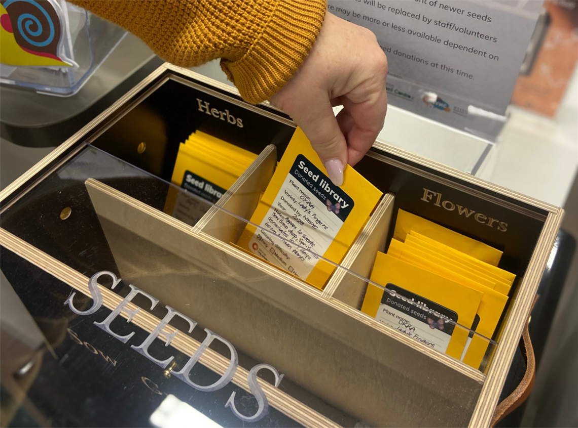 A photo of a person wearing a mustard-coloured jumper's hand lifting a seed packet from a wooden box at the seed library.