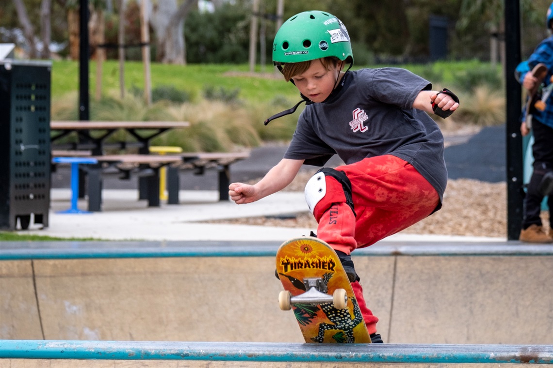 A young boy with a green helmet, black T-shirt and orange pants stalls on his skateboard atop a ramp.
