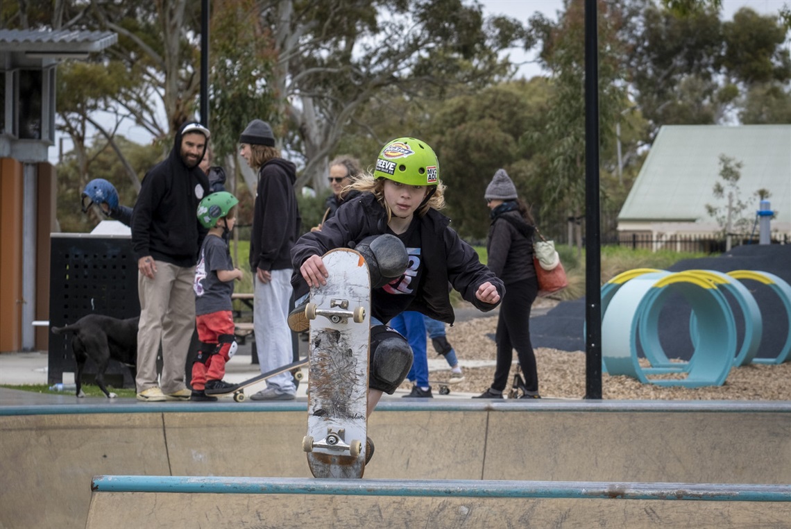 A young skateboarder in a green helmet launches out of a skateboard park bowl.